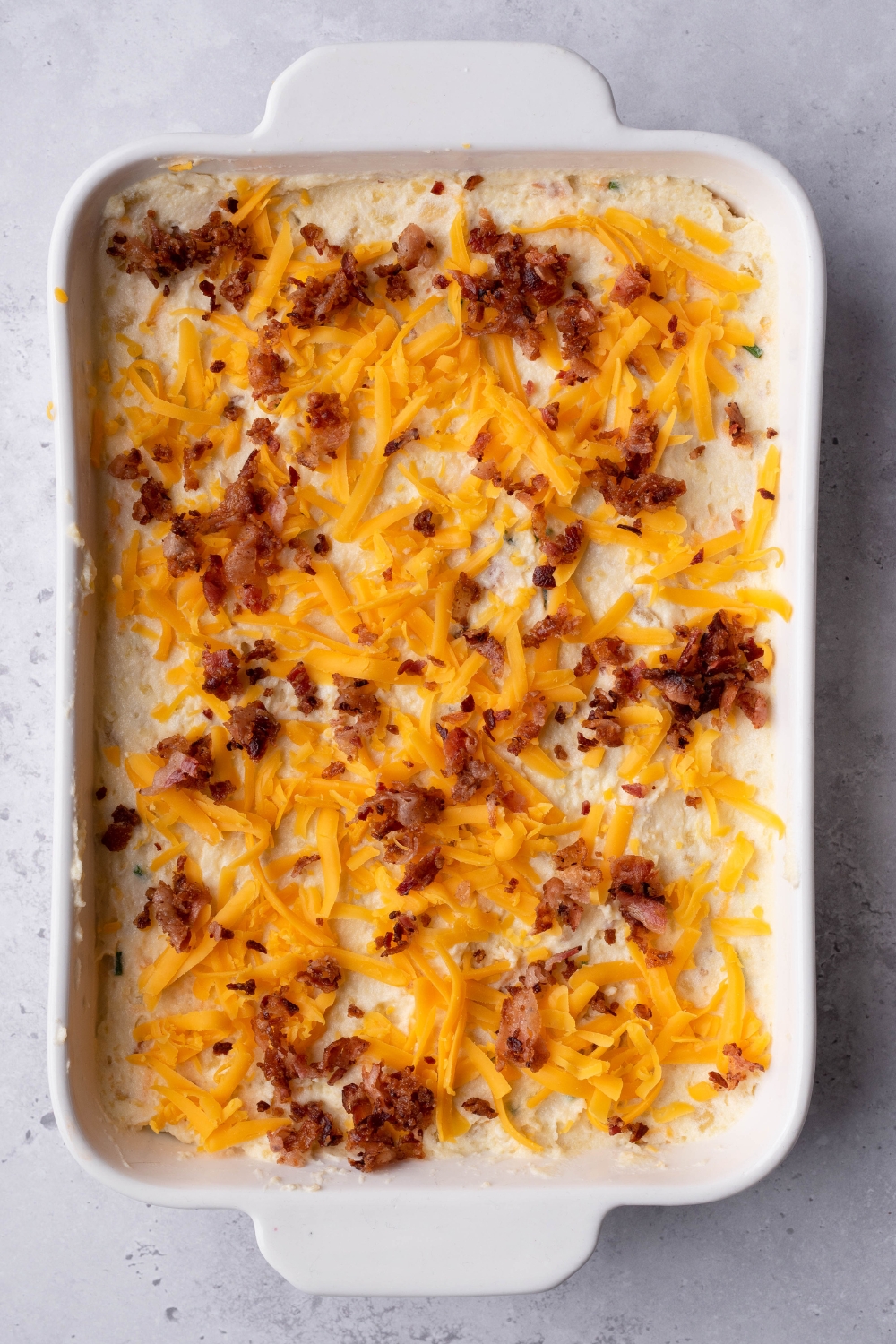 A baking casserole dish with unbaked potato casserole topped with extra bacon bits and shredded cheese on top.
