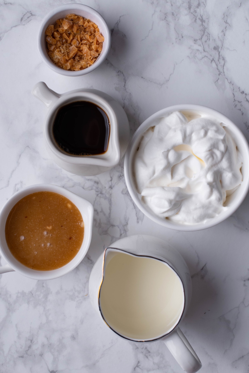 A countertop with a small bowl of whipped cream, a small bowl of caramel sauce, a pitcher of coffee, a small bowl of crumbly topping, and a small pitcher of milk.