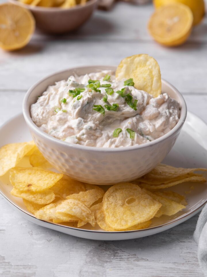 A small dip dish with clam dip in it being served on a plate with potato chips.\