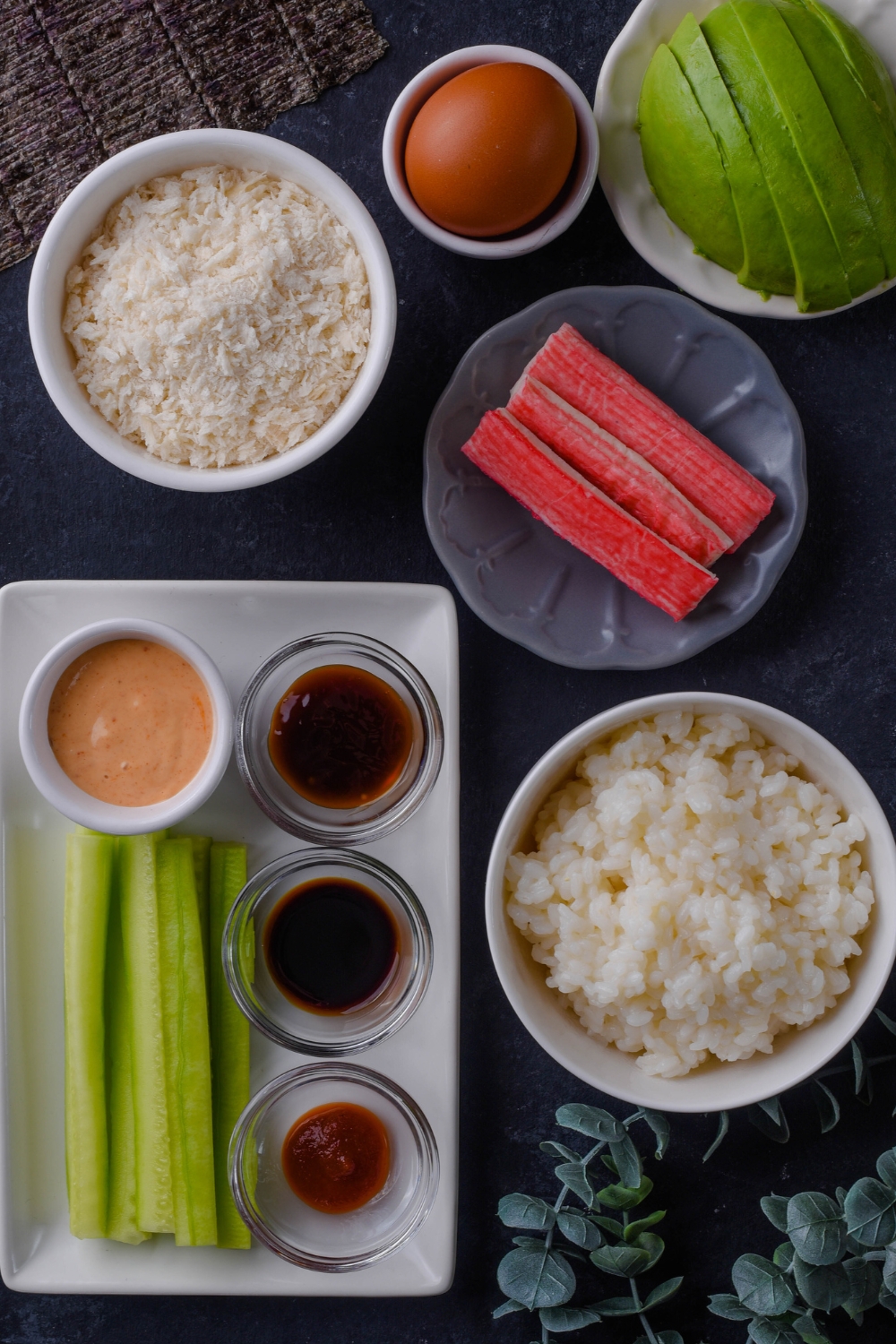 An overhead view of cooked rice, imitation crab, Half of an avocado, an egg, breadcrumbs, spicy mayo, soy sauce, oyster sauce, Sriracha sauce, and sliced cucumbers.
