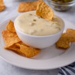 A small bowl with queso served on a plate with tortilla chips. A tortilla chip is dipped in the queso.