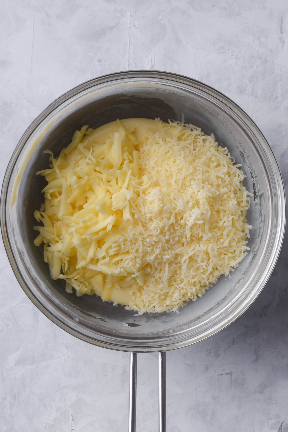 A double boiler with shredded cheeses being added to melted white american cheese mix.