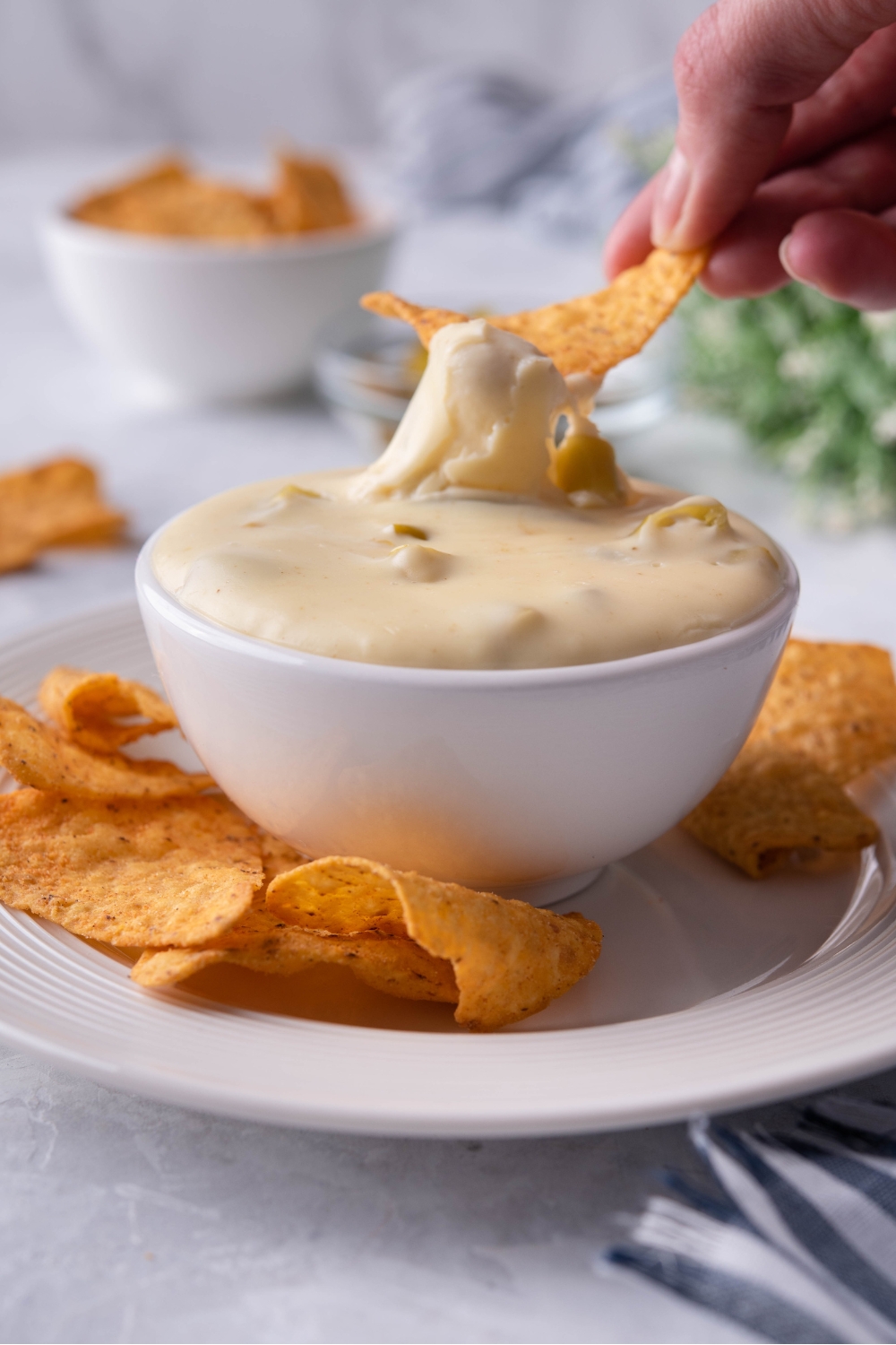 A small bowl with queso on a plate with tortilla chips. A hand is dipping a tortilla chip into the queso.