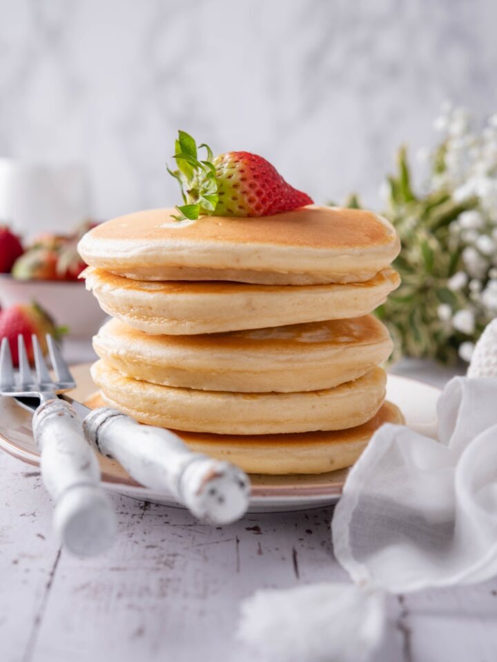 A stack of fluffy pancakes covered in maple syrup with a strawberry on top. The pancakes are on a white plate with a set of silverware.