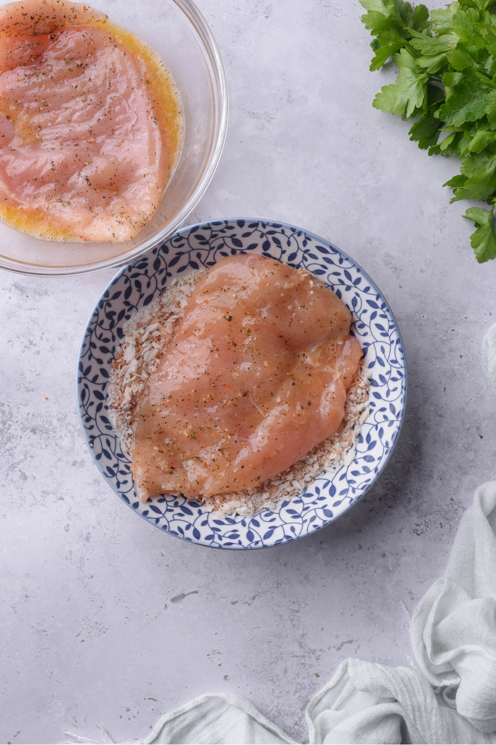 A blue and white bowl with a seasoned chicken cutlet in it. Next to the bowl is a second bowl of a marinating chicken cutlet.