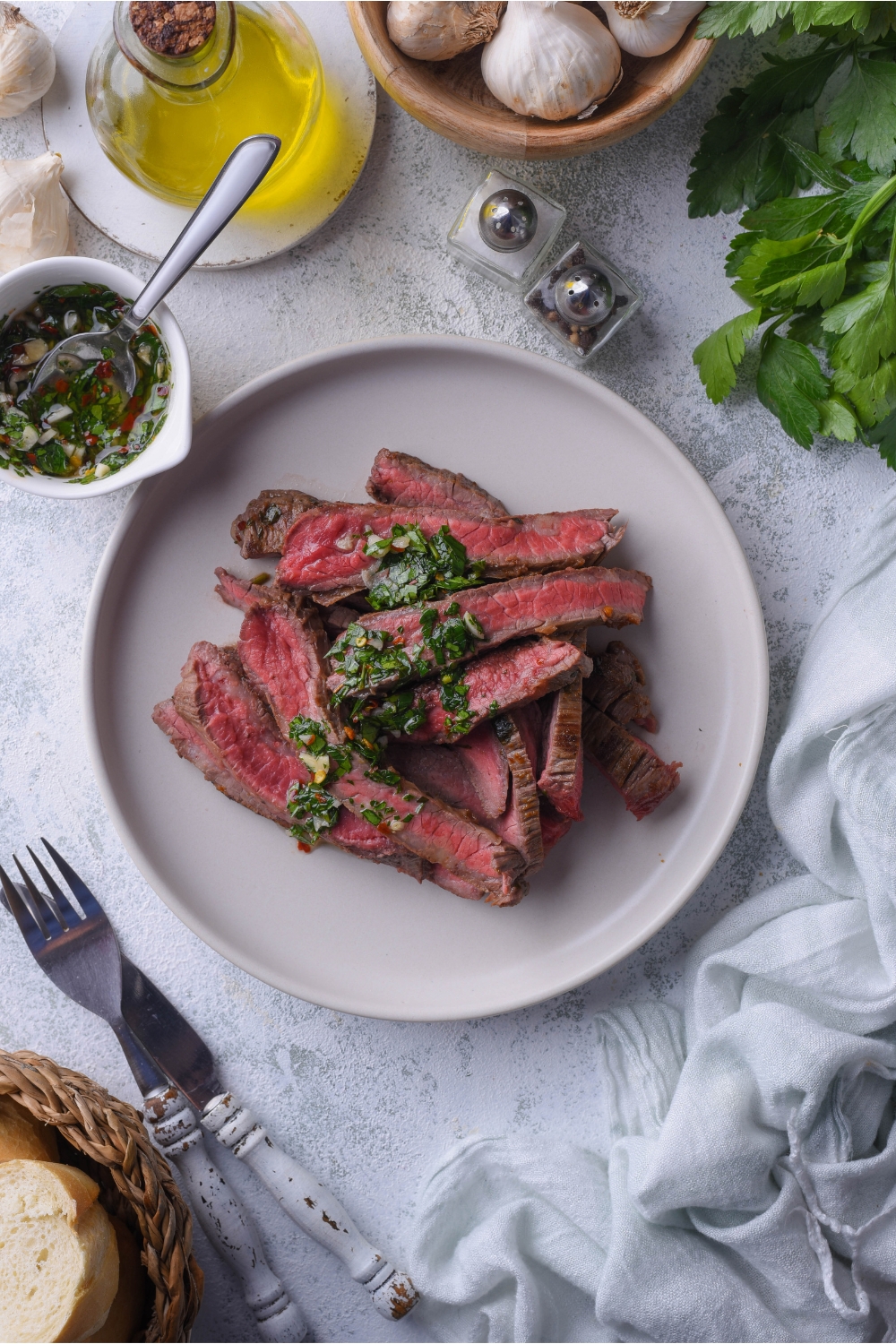 Thinly sliced bavette steak piled on a white plate and covered in chimichurri sauce. The plate of steak is surrounded by an assortment of ingredients.