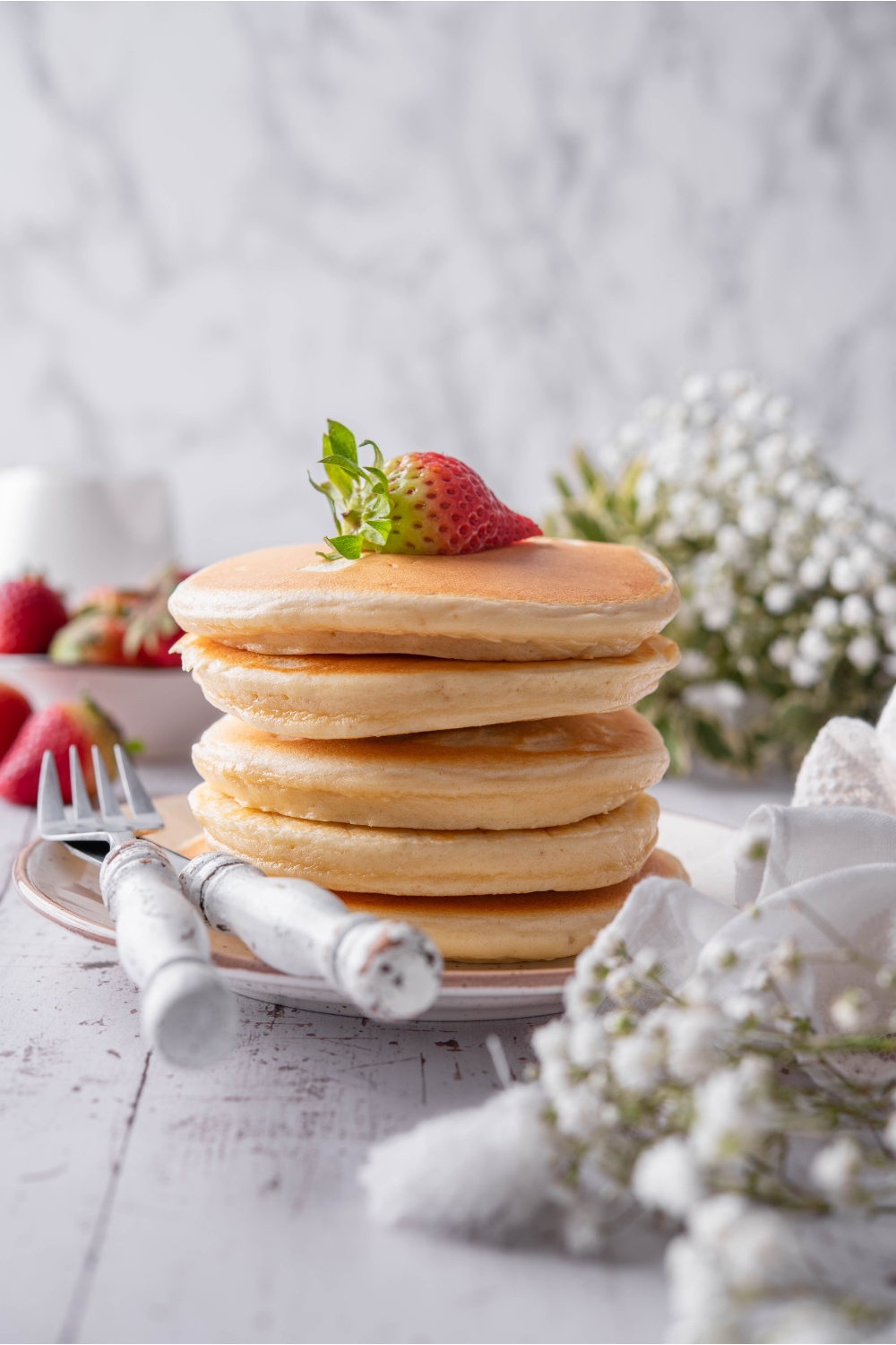 A stack of fluffy pancakes covered in maple syrup with a strawberry on top. The pancakes are on a white plate with a set of silverware.