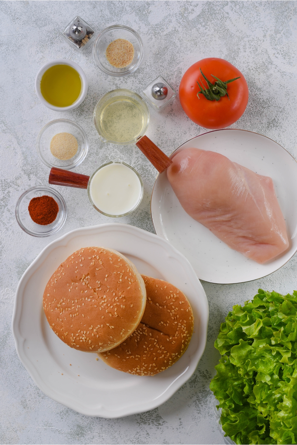 Overhead view of an assortment of ingredients including a plate of hamburger buns, a plate of raw chicken, a large tomato, and bowls of spices, oil, and buttermilk.