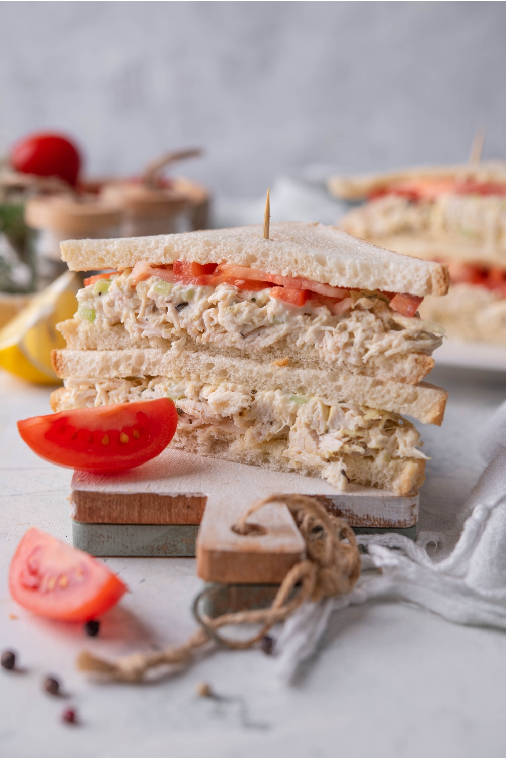 A chicken salad sandwich with tomato cut in half with both halves on top of each other on a serving board with a tomato wedge next to it.