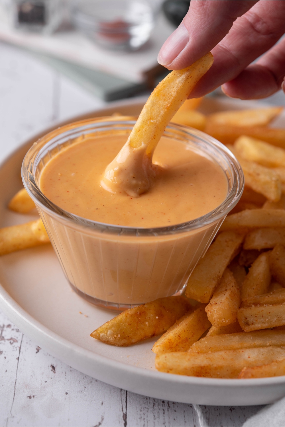 A hand dipping a french fry into a clear bowl of melted cheese sauce. The bowl of sauce is on a plate filled with seasoned french fries.