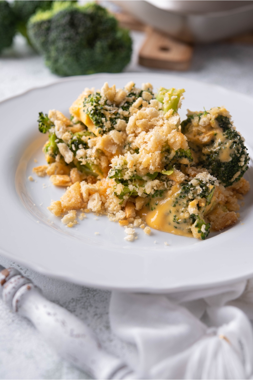 A serving of Velveeta broccoli casserole on a white plate with silverware next to the plate and broccoli florets in the background.