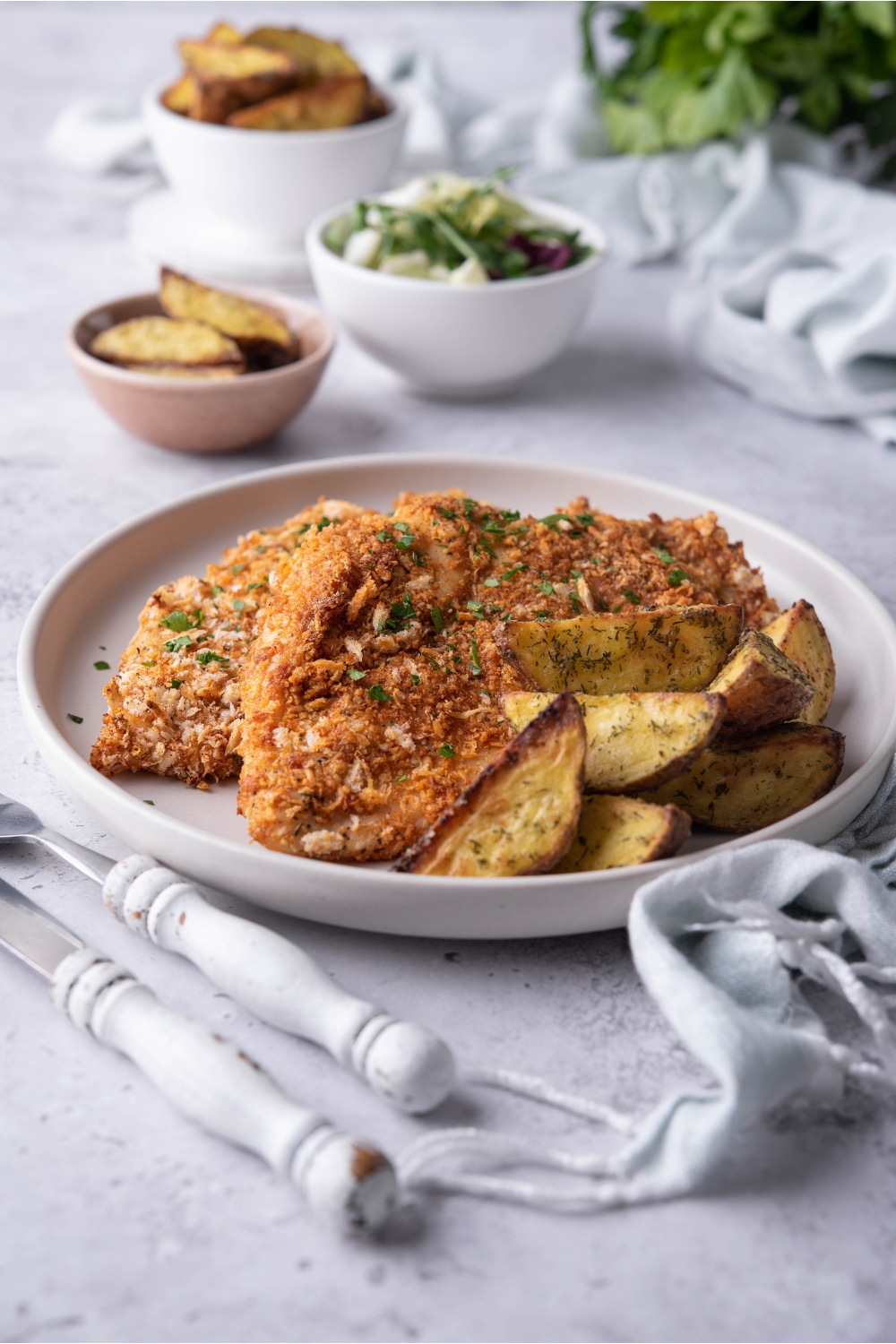 A plate of seasoned baked chicken cutlets garnished with fresh herbs and served with a side of potato wedges. There's a set of silverware next to the plate.