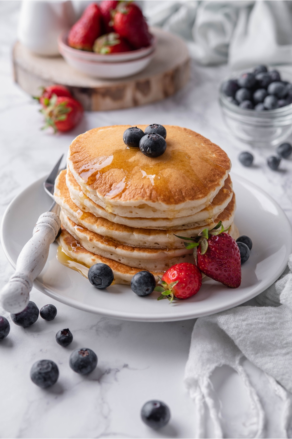 A stack of pancakes topped with maple syrup and blueberries on a white plate with a fork and strawberries on the plate beside the pancakes. Berries are scattered on the counter and in the background.