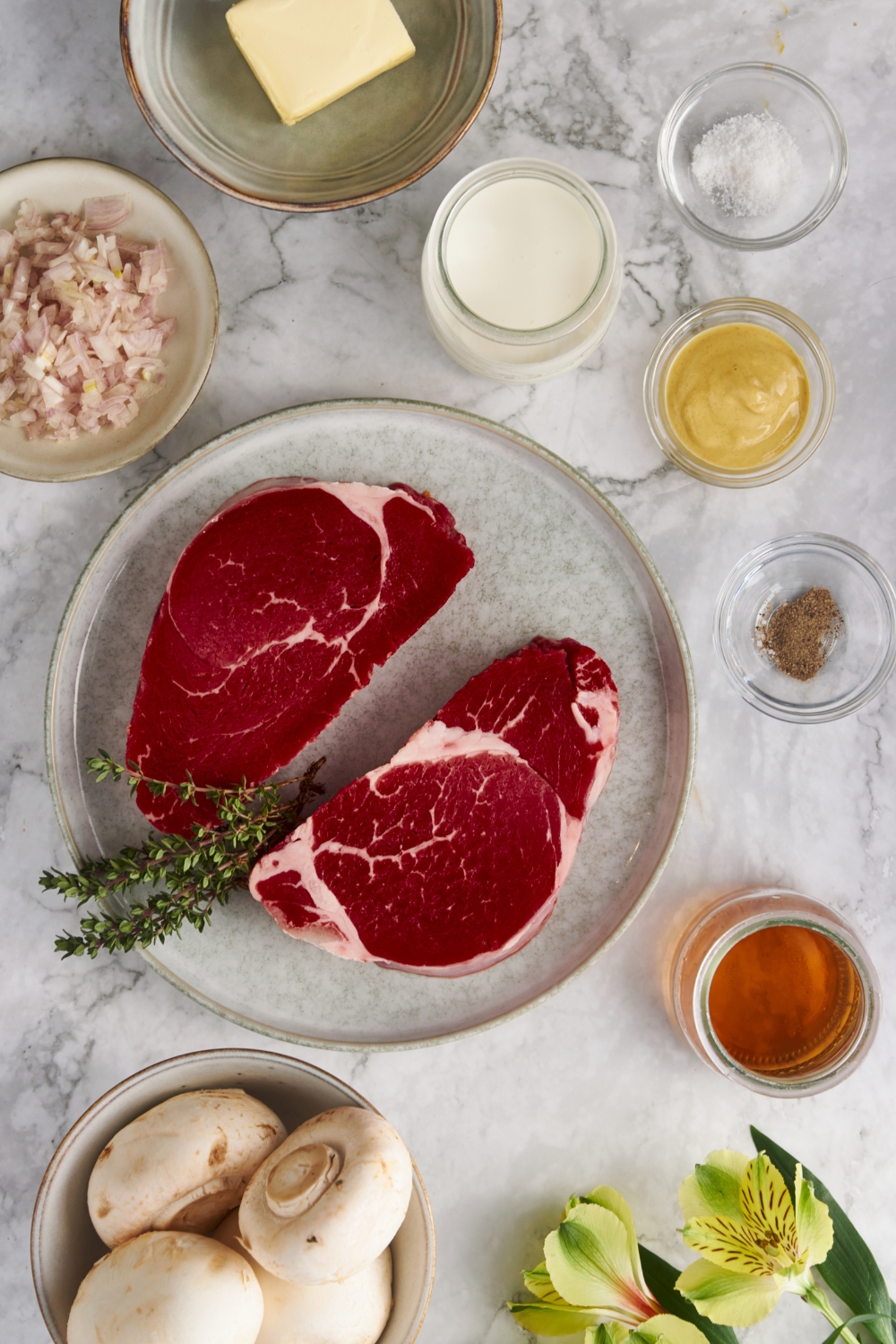 Overhead view of an assortment of ingredients including two raw steaks, fresh herbs, jars of brandy and cream, and bowls of salt, pepper, mustard, mushrooms, butter, and diced shallots.