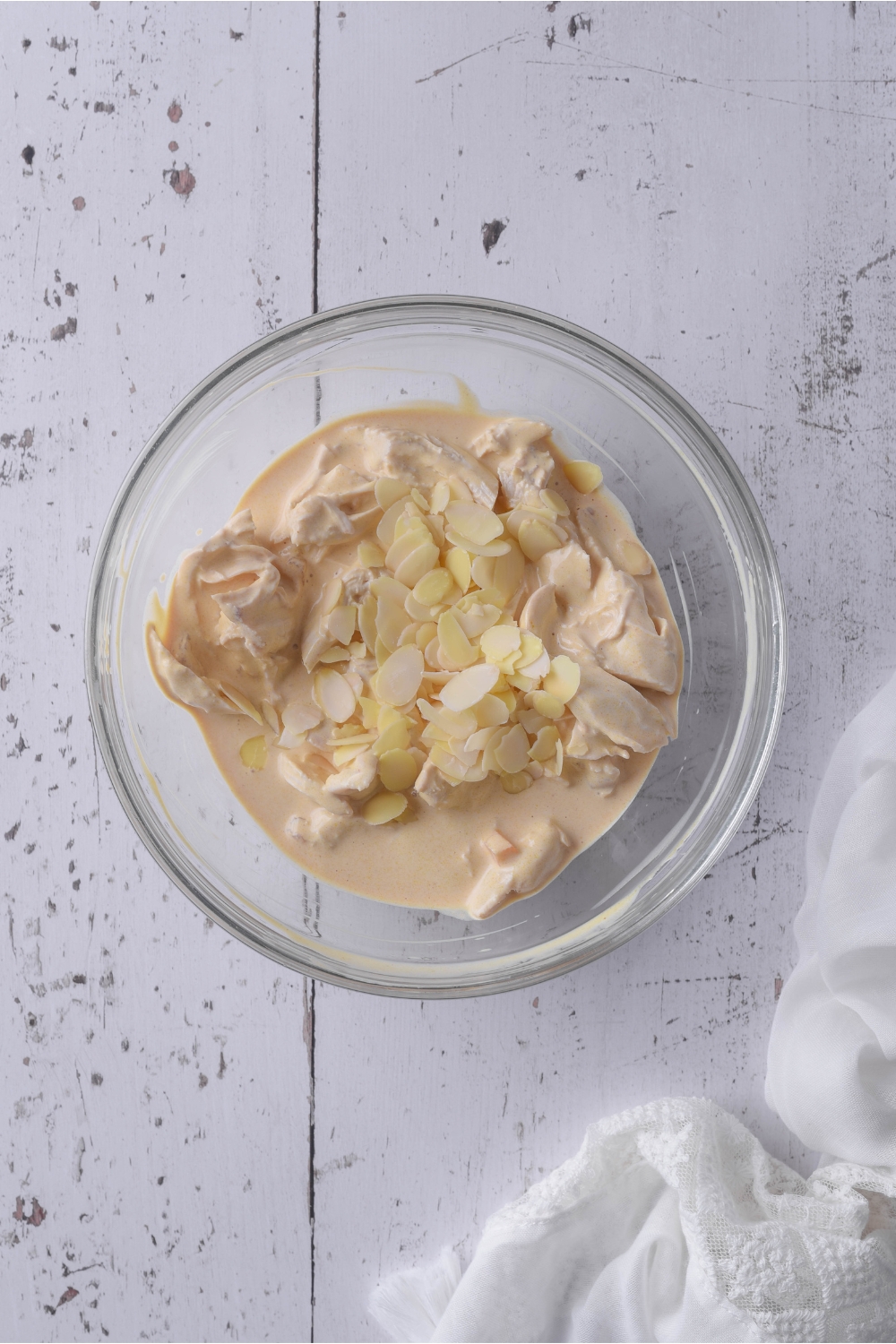 A clear bowl filled with cooked and shredded chicken in a creamy sauce and slivered almonds atop the chicken.