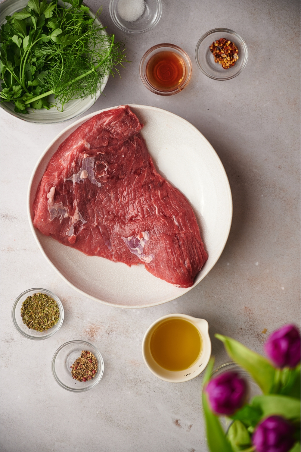 A raw tri-tip steak surrounded by ingredients including bowls of fresh herbs, red wine vinegar, red pepper flakes, black pepper, and oil.
