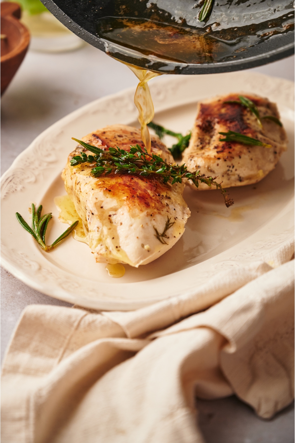 Two cooked chicken breasts topped with fresh herbs and a skillet pouring a butter sauce overtop of one of the chicken breasts.