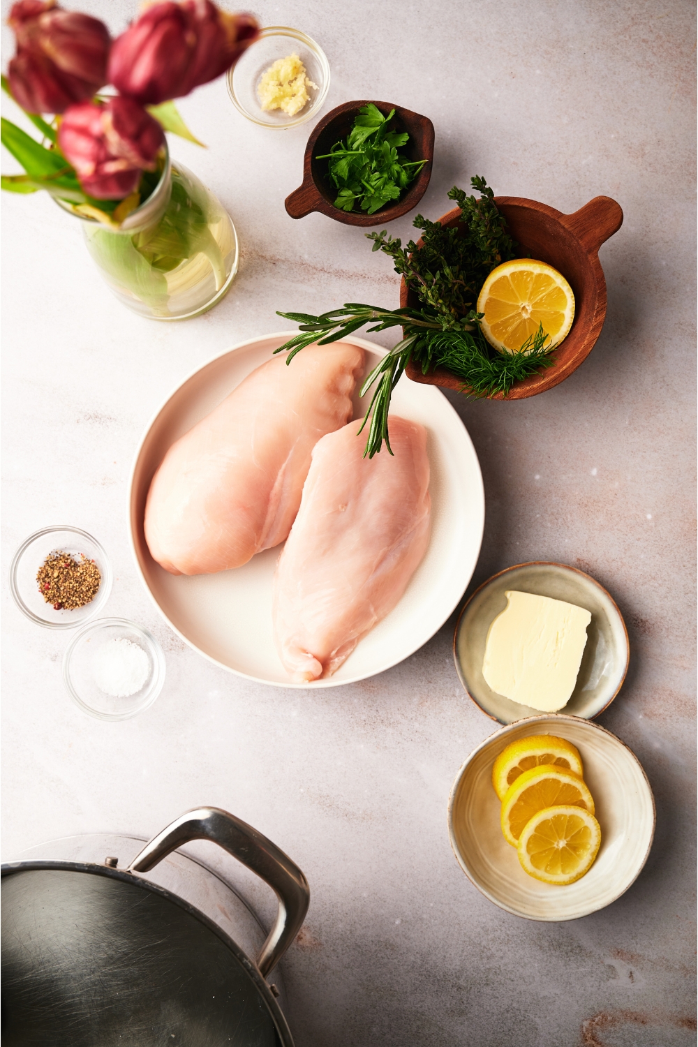 An assortment of ingredients including a plate of raw chicken and bowls of salt, pepper, butter, lemon slices, and fresh herbs.