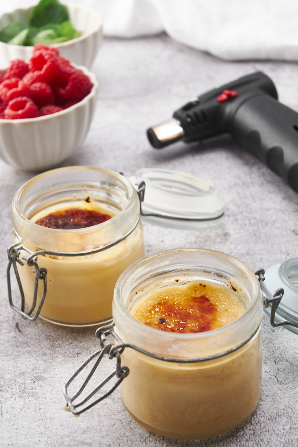 Two creme brûlée desserts in jars with a kitchen torch and a bowl of raspberries in the background.