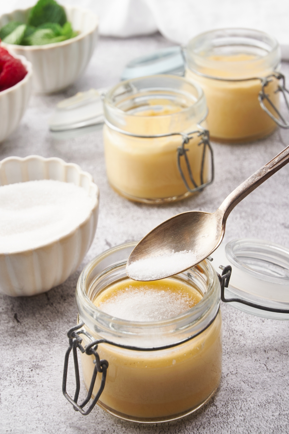 A spoon spooning sugar on top of a cooked jar of custard, with two more jars of custard in the background.