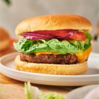 A cheeseburger with red onion, lettuce, and tomato on it. The burger is on a white plate atop a wood cutting board.