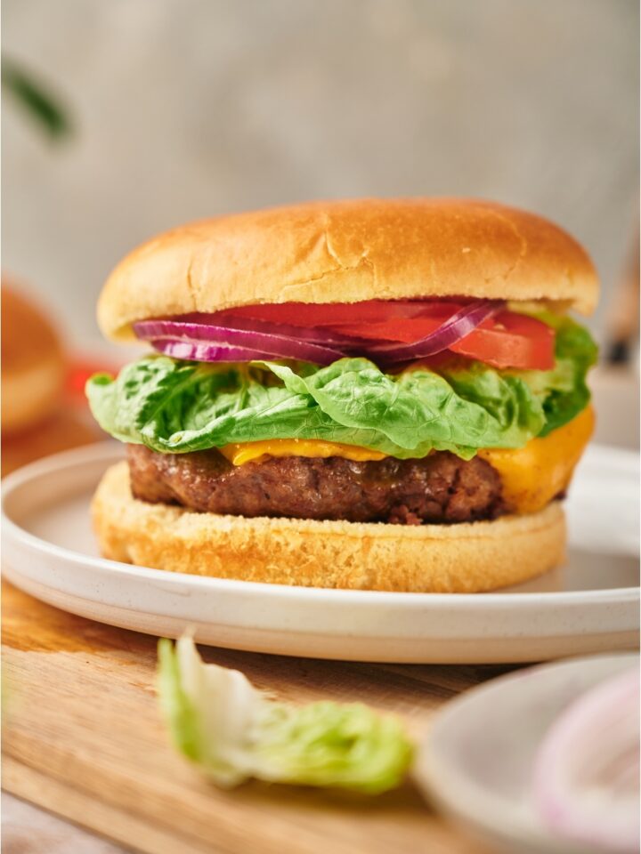 A cheeseburger with red onion, lettuce, and tomato on it. The burger is on a white plate atop a wood cutting board.