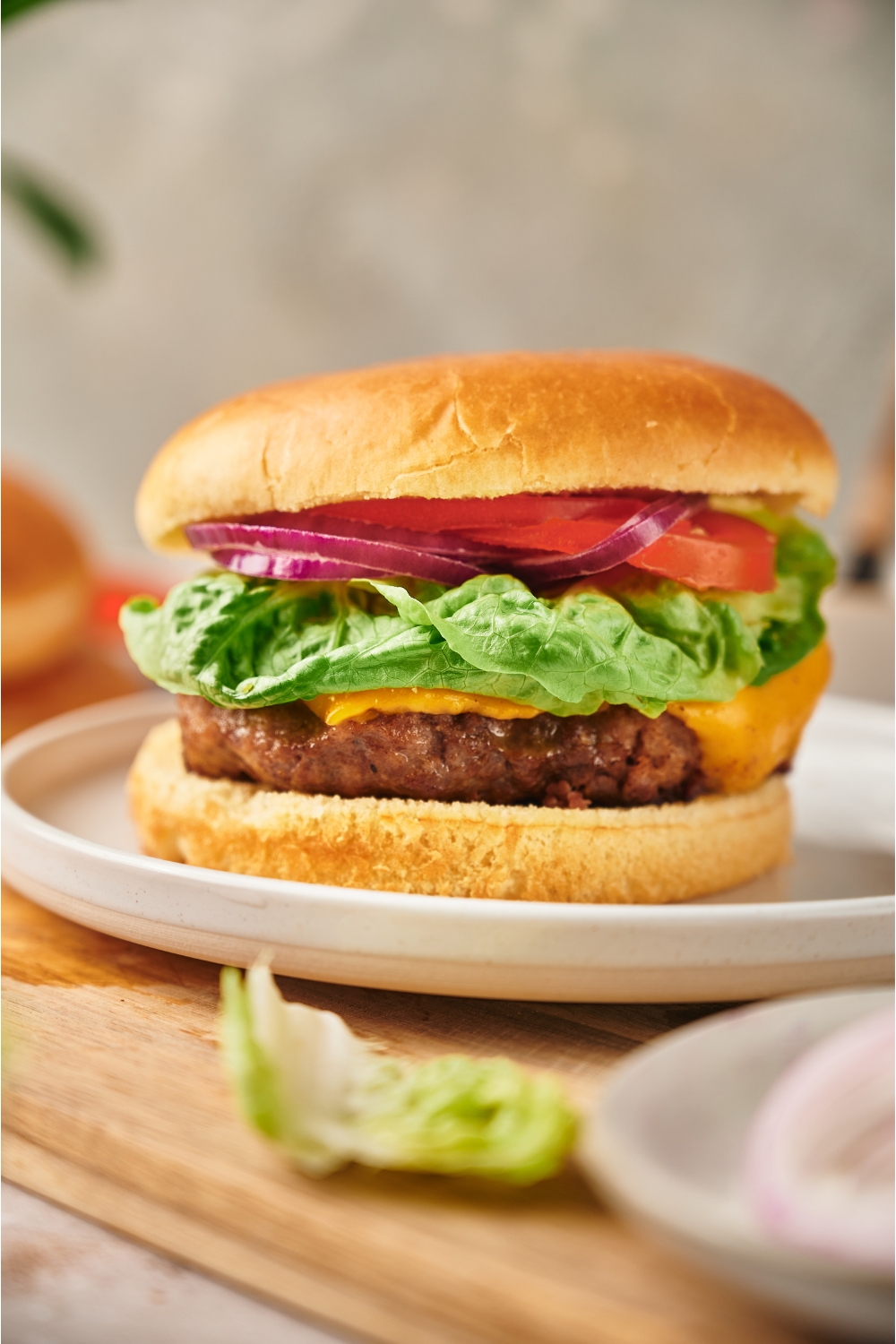 A cheeseburger with red onion, lettuce, and tomato on it. The burger is on a white plate atop a wood cutting board.