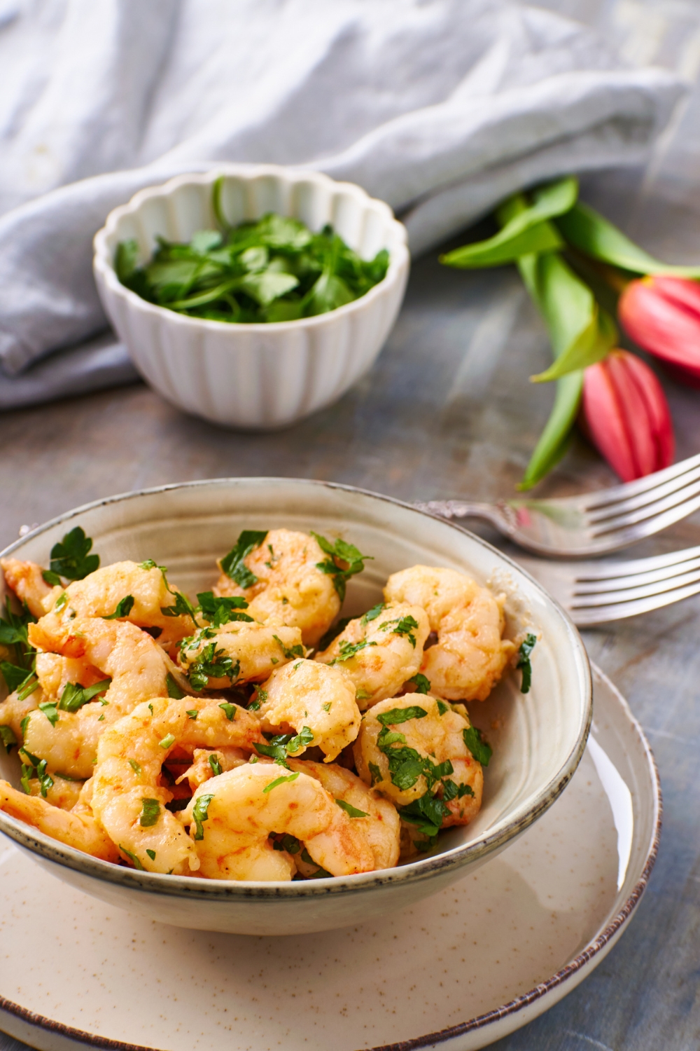 Cooked shrimp garnished with fresh herbs in a bowl. The bowl is on top of a plate and two forks are next to it.
