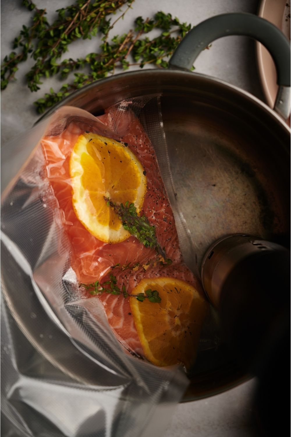 Salmon loins in a vacuum-sealed bag with orange slices and fresh herbs. The salmon is in a large water bath with a sous vide machine in the pot.