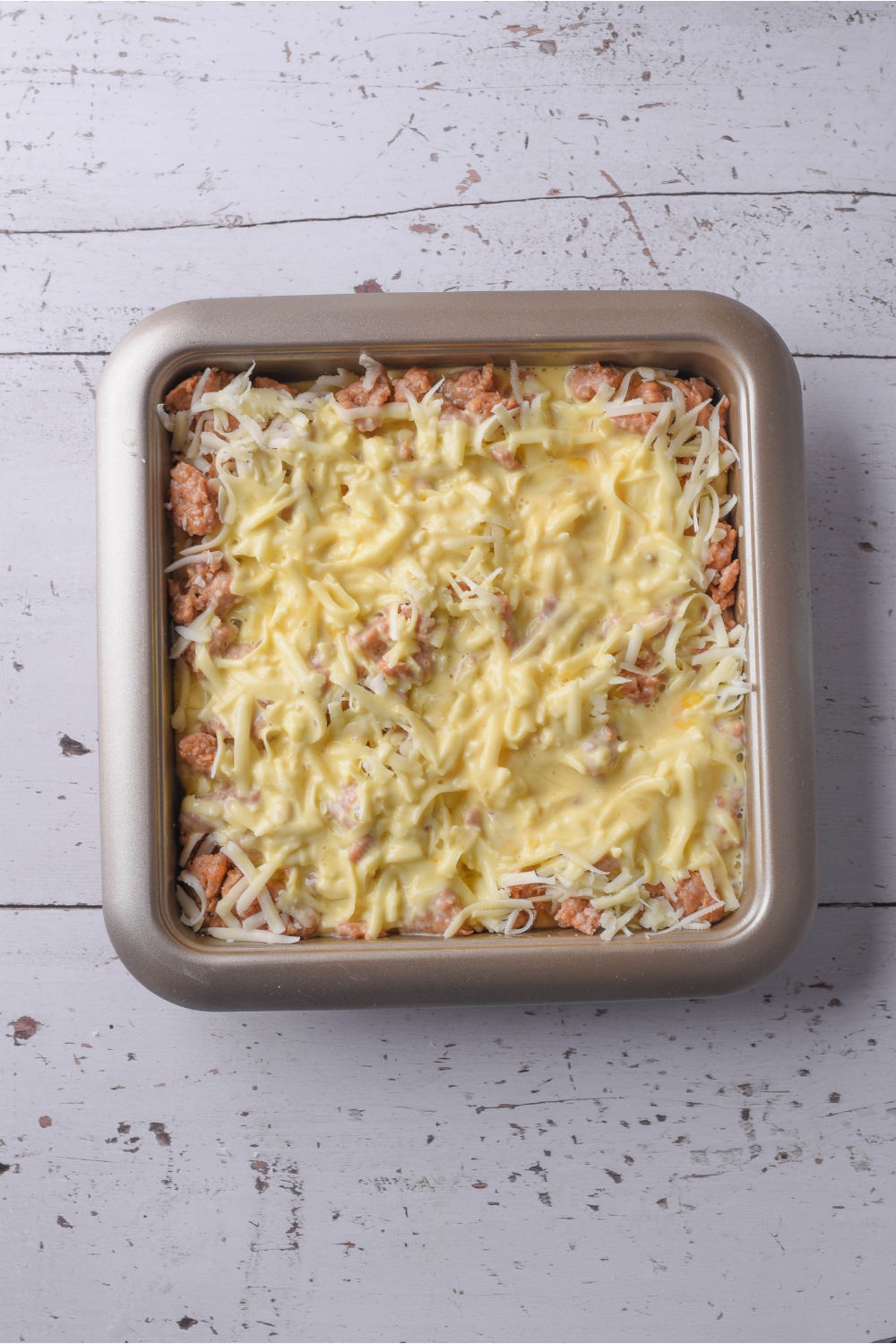 A square baking dish filled with cooked sausage, shredded cheese, and a beaten egg and milk mixture.