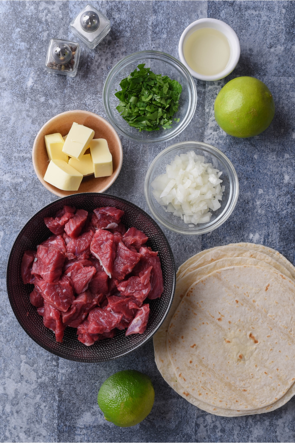Overhead view of an assortment of ingredients including bowls of raw ground beef, diced onions, butter, chopped cilantro, and a stack of tortillas.