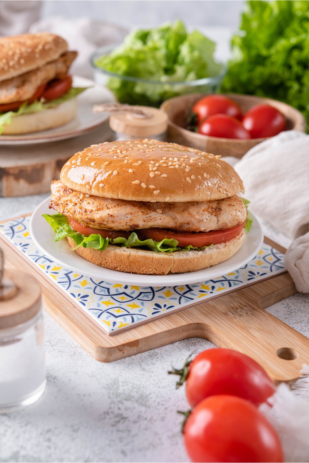 A grilled chicken sandwich with lettuce and sliced tomatoes on a sesame seed bun. The sandwich is on a white plate atop a decorative serving tray and there is a second sandwich in the background.