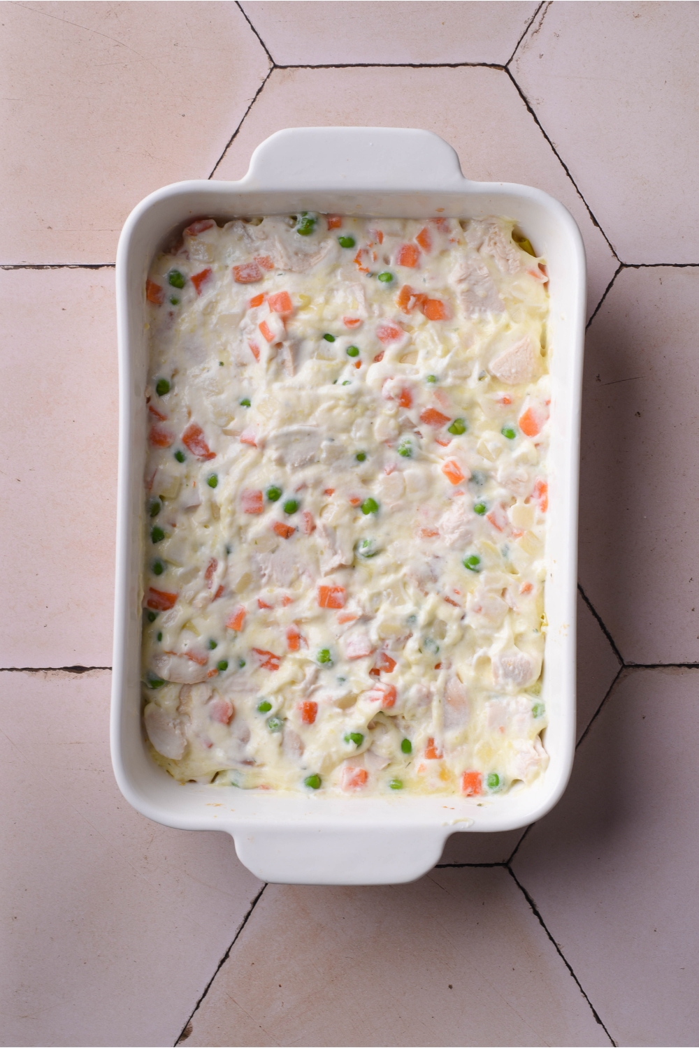A white baking dish filled with a mixture of creamy soup, diced carrots, peas, and diced chicken.