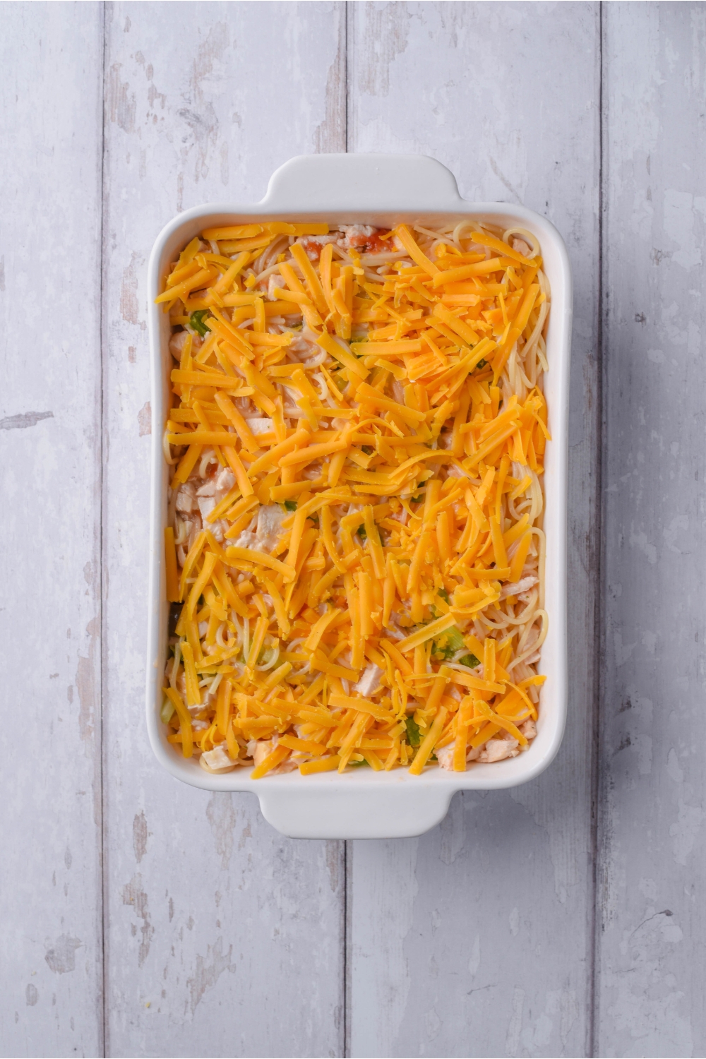 An unbaked chicken spaghetti casserole covered in shredded cheese in a white baking dish.