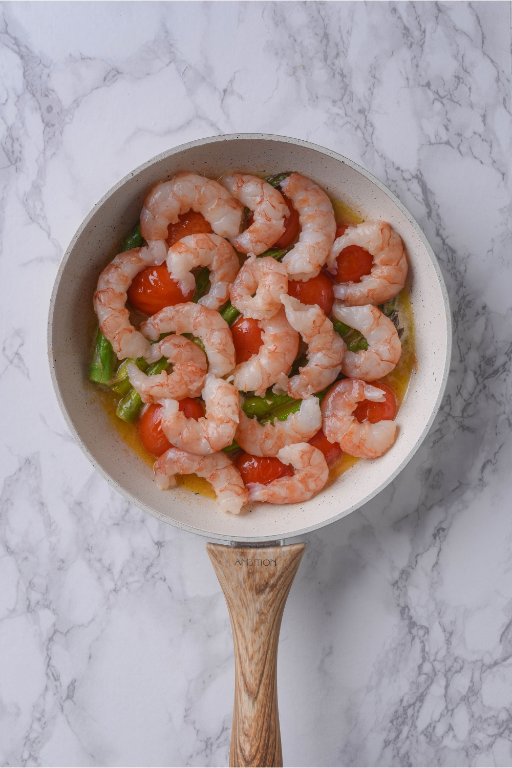 A grey skillet filled with melted butter, asparagus, cherry tomatoes, and raw shrimp on top.