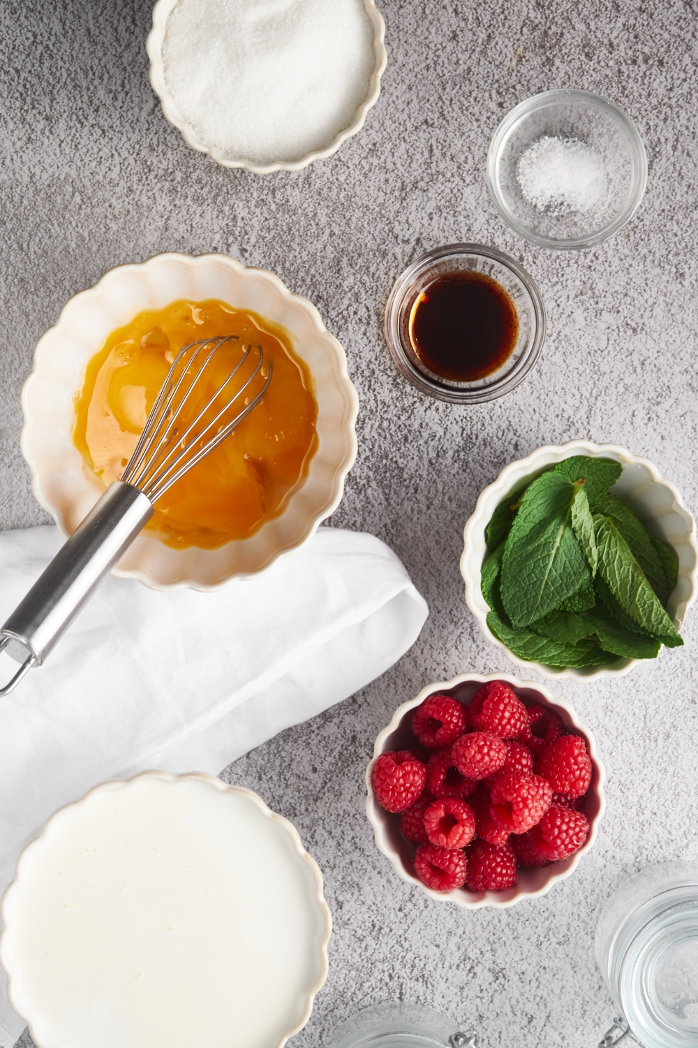 Overhead view of an assortment of ingredients including bowls of mint leaves, raspberries, vanilla extract, whisked eggs, and milk.