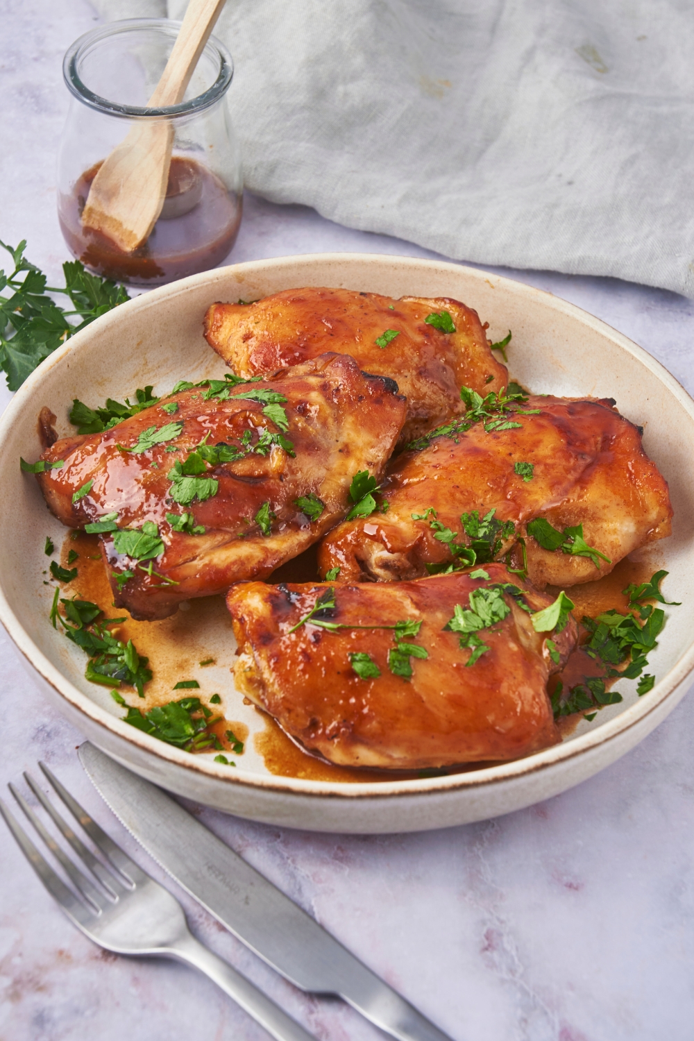 Baked chicken thighs in on a white plate. The chicken is coated in a brown sauce and garnished with fresh herbs and there is a fork and knife next to the chicken.