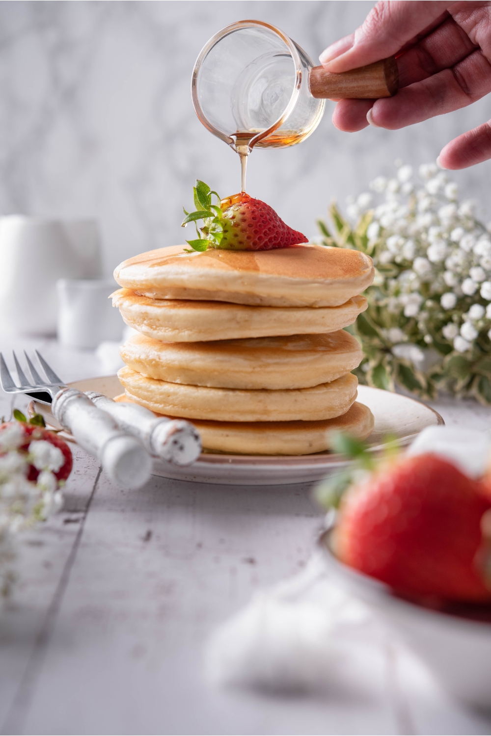 A hand pouring maple syrup over a giant stack of fluffy pancakes. The pancakes have a single strawberry on top and a set of silverware next to them.