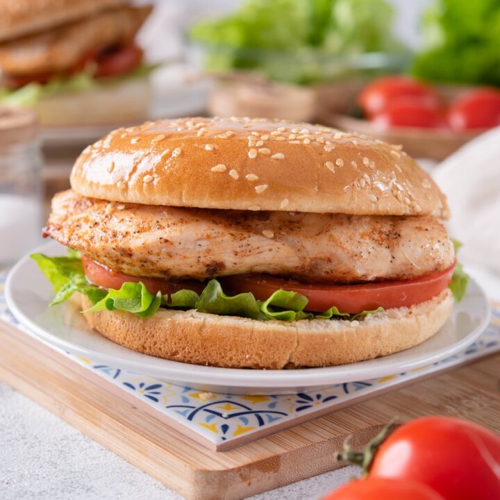 A grilled chicken sandwich with lettuce and sliced tomatoes on a sesame seed bun. The sandwich is on a white plate atop a decorative serving tray and there is a second sandwich in the background.