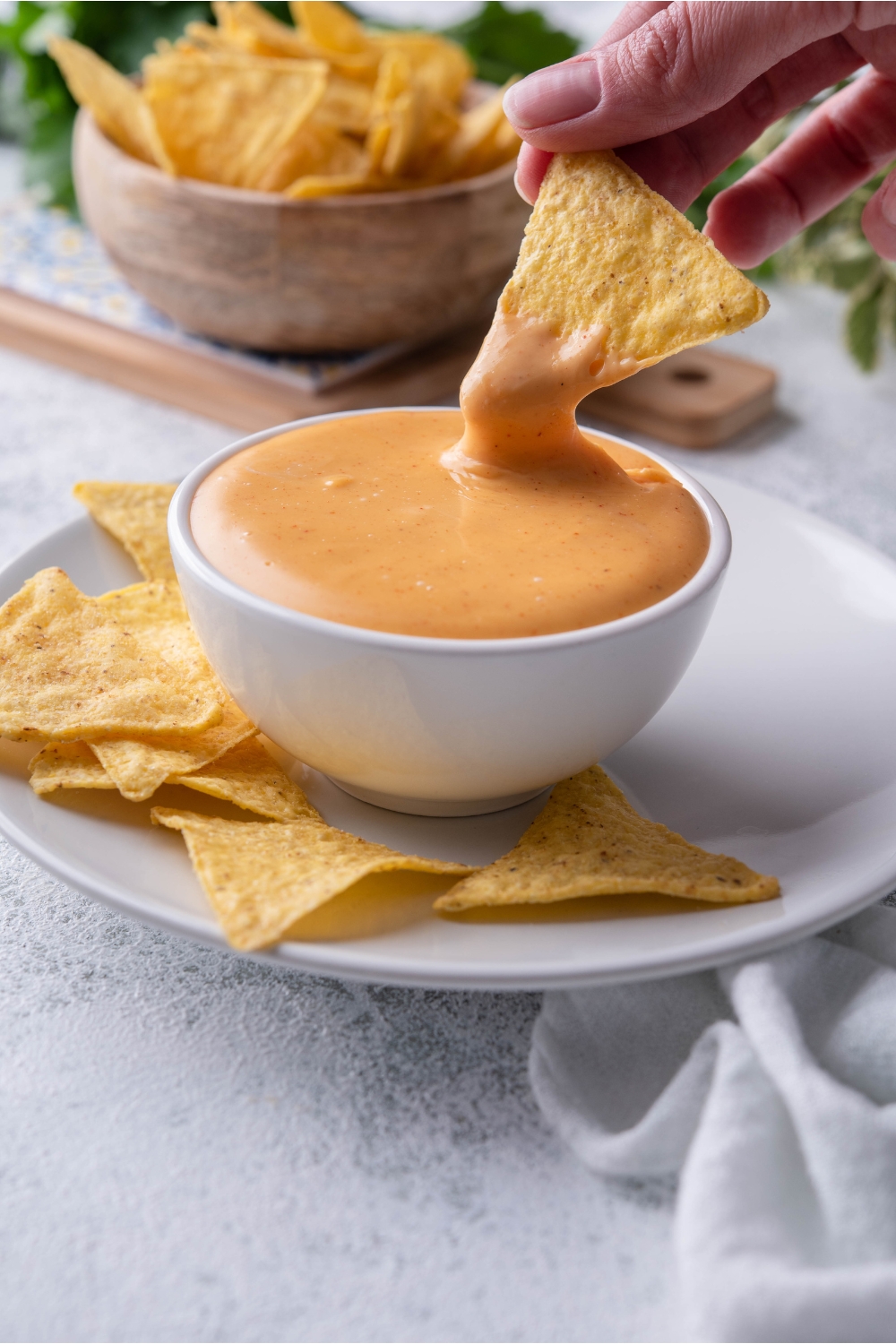 A hand dipping a tortilla chip into a bowl of Taco Bell nacho cheese sauce. The cheese sauce is in a white bowl atop a white plate filled with more tortilla chips.