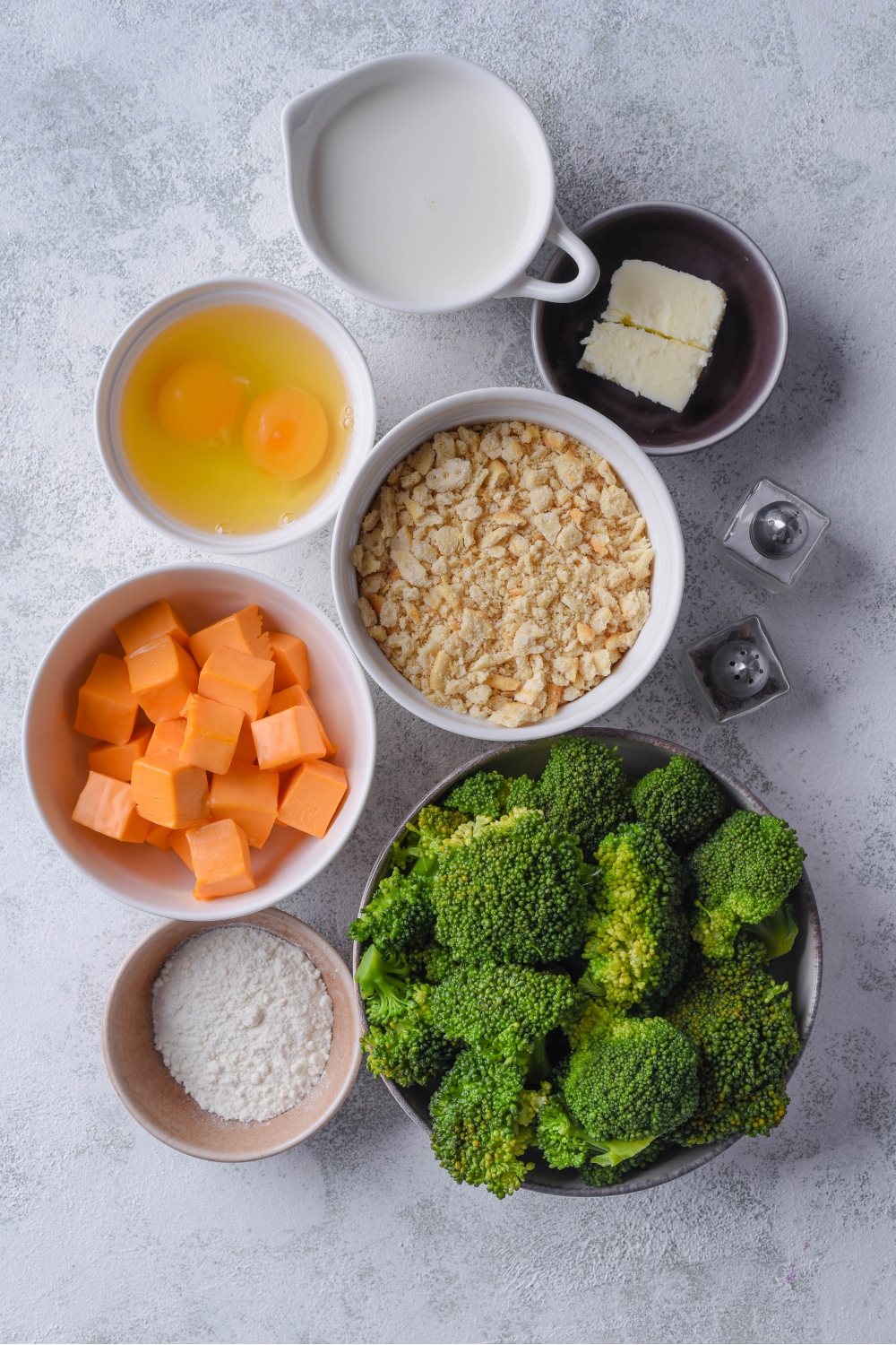 An assortment of ingredients including bowls of raw eggs, cubed cheese, cracker crumbs, milk, butter, broccoli, and flour.
