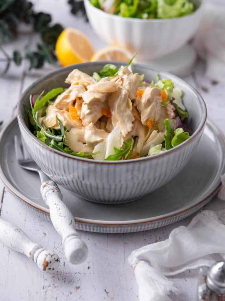 A blue bowl filled with coronation chicken salad atop a bed of greens. The bowl is on a blue plate and there is a fork on the plate.