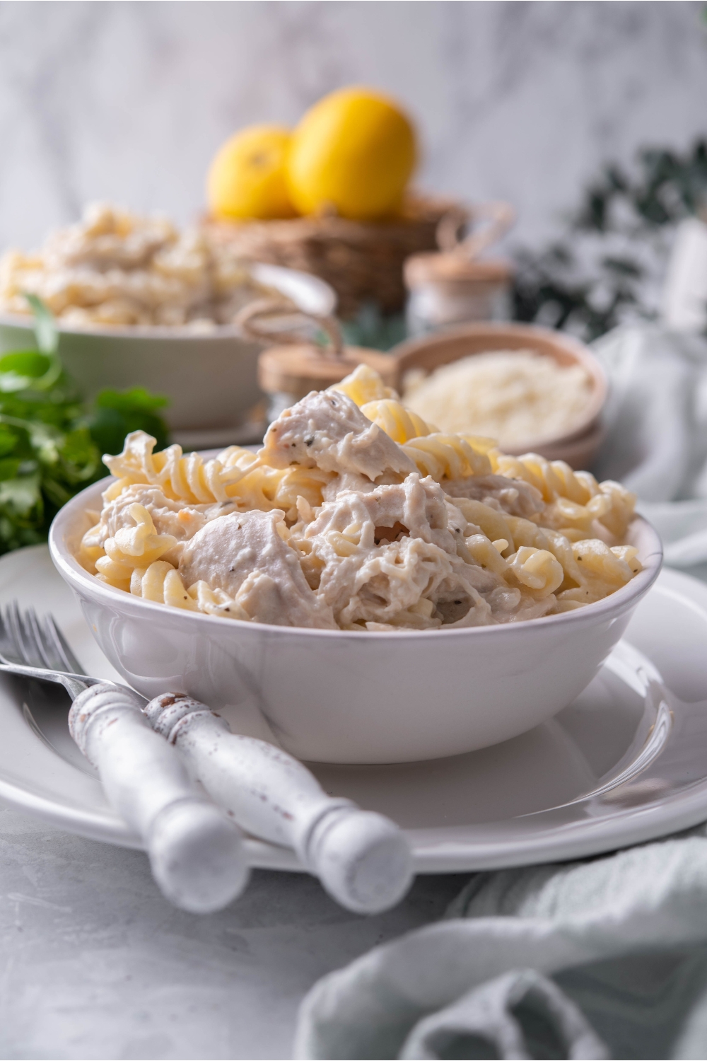 A bowl of pasta and chicken in a cream sauce atop a white plate with two forks next to the bowl.