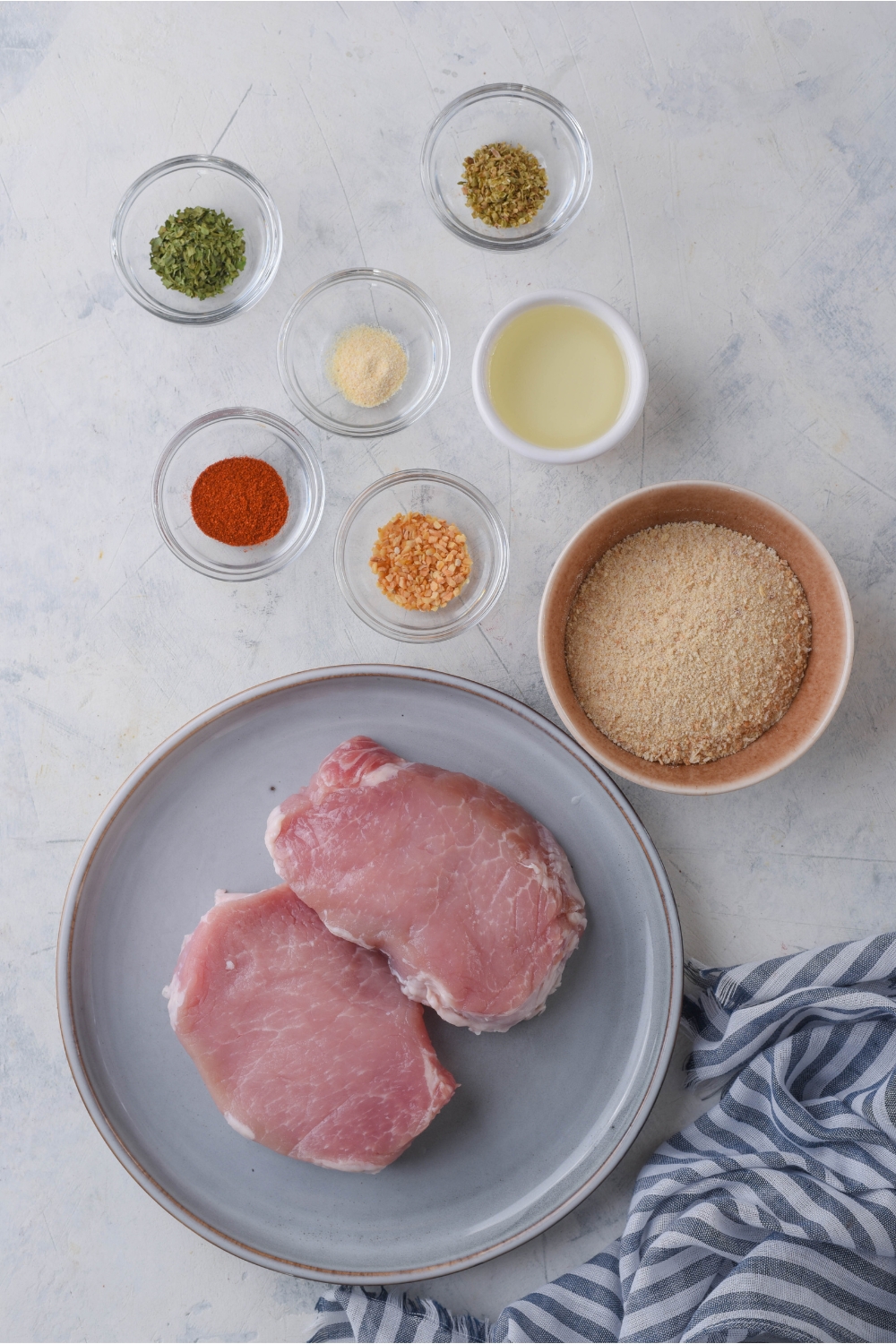 An assortment of ingredients including a plate of pork chops, bread crumbs, and spices.