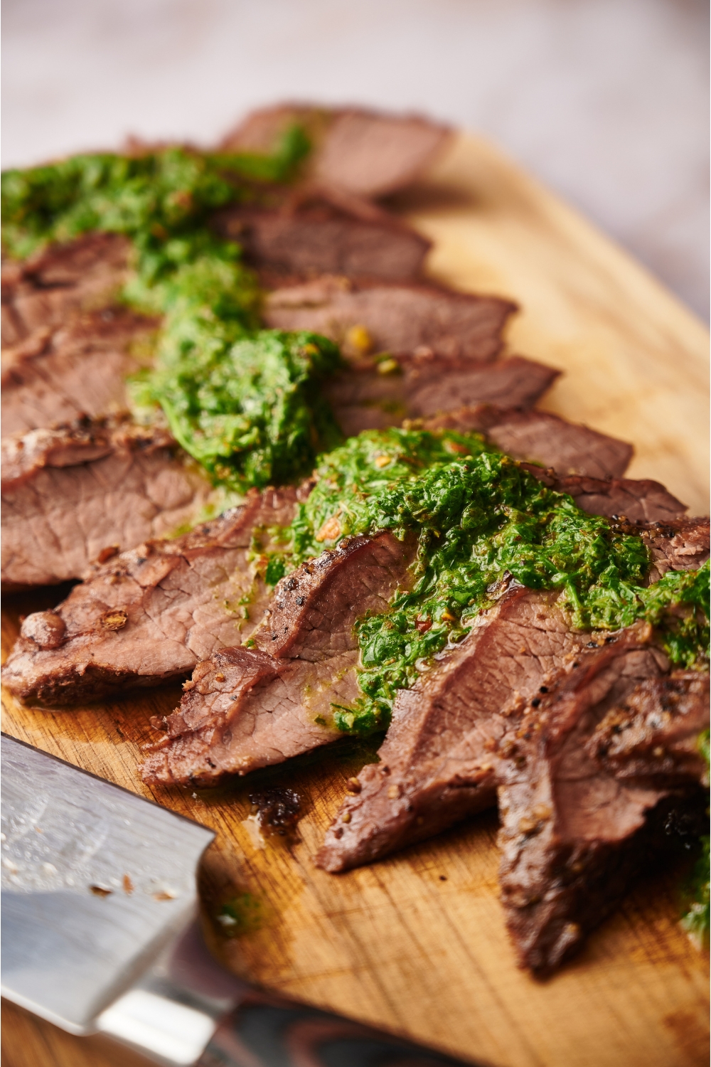 Close up of thinly sliced steak on a wooden board that's been covered in chimichurri sauce.