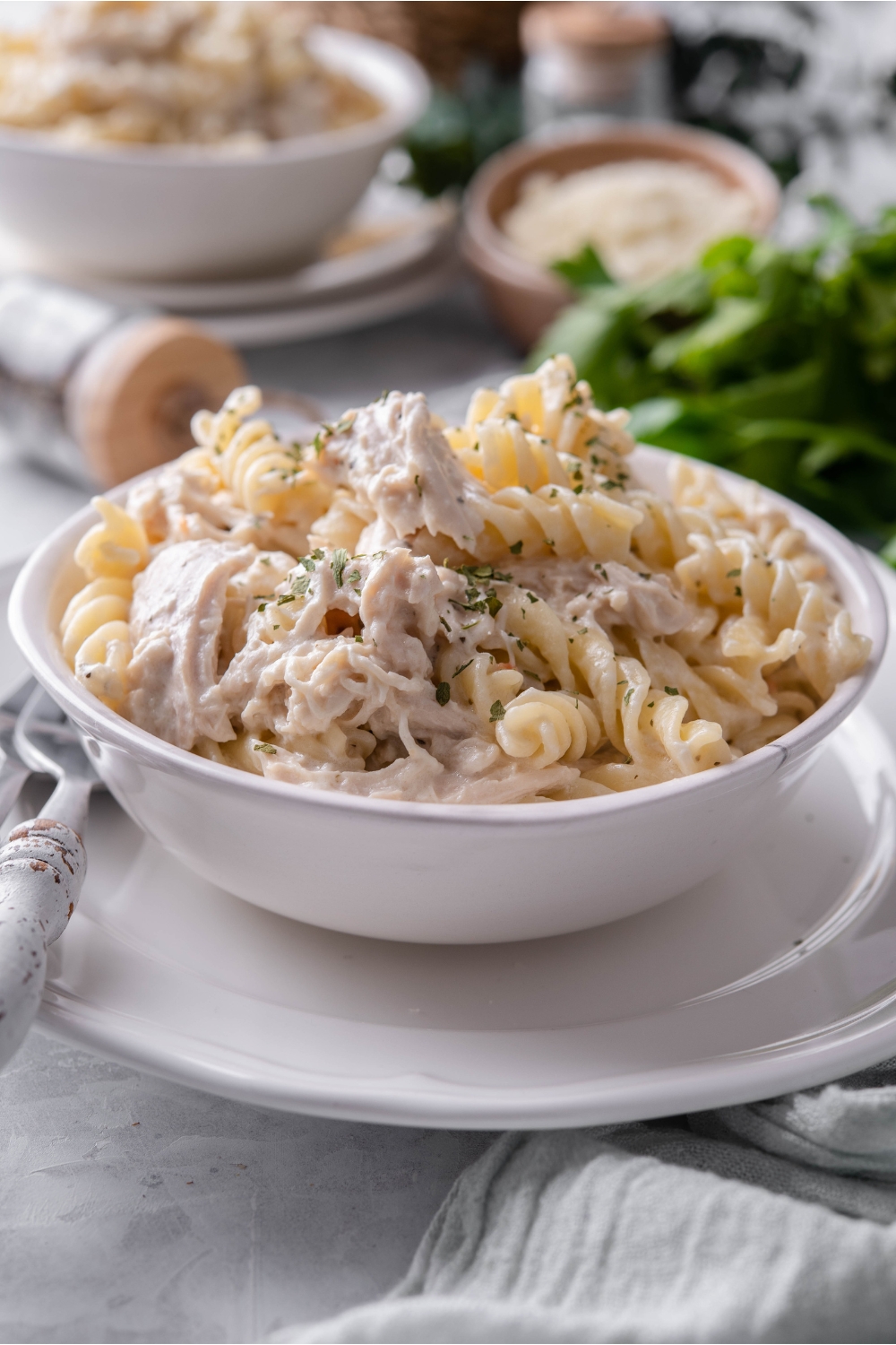 A bowl of pasta and chicken in a cream sauce atop a white plate with a fork next to the bowl and a second bowl of pasta in the background.