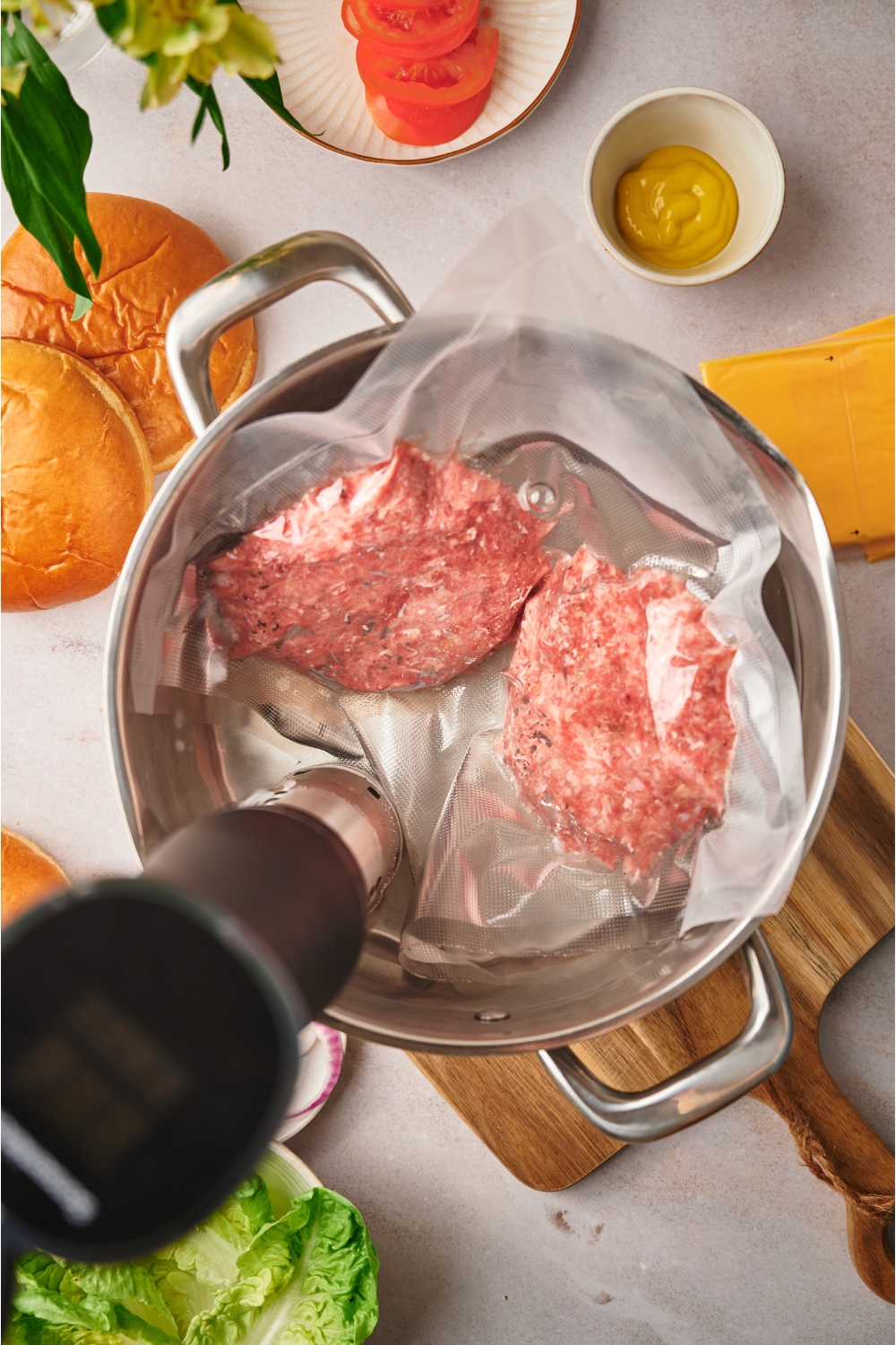 Two burger patties in a sous vide bag in a water bath with a sous vide machine in it. The water bath is surrounded by ingredients for making cheeseburgers.