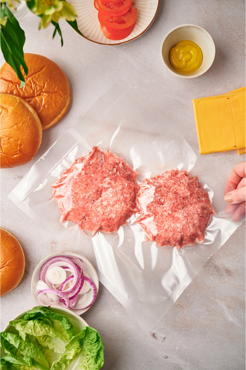 A hand holding a sous vide bag with two uncooked burger patties in it. The bag is surrounded by ingredients for making cheeseburgers.