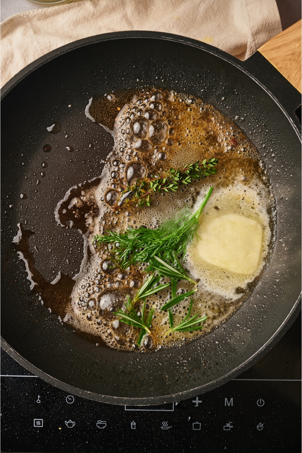 A black skillet with melting butter and fresh green herbs in it. The butter is bubbling and almost completely melted.