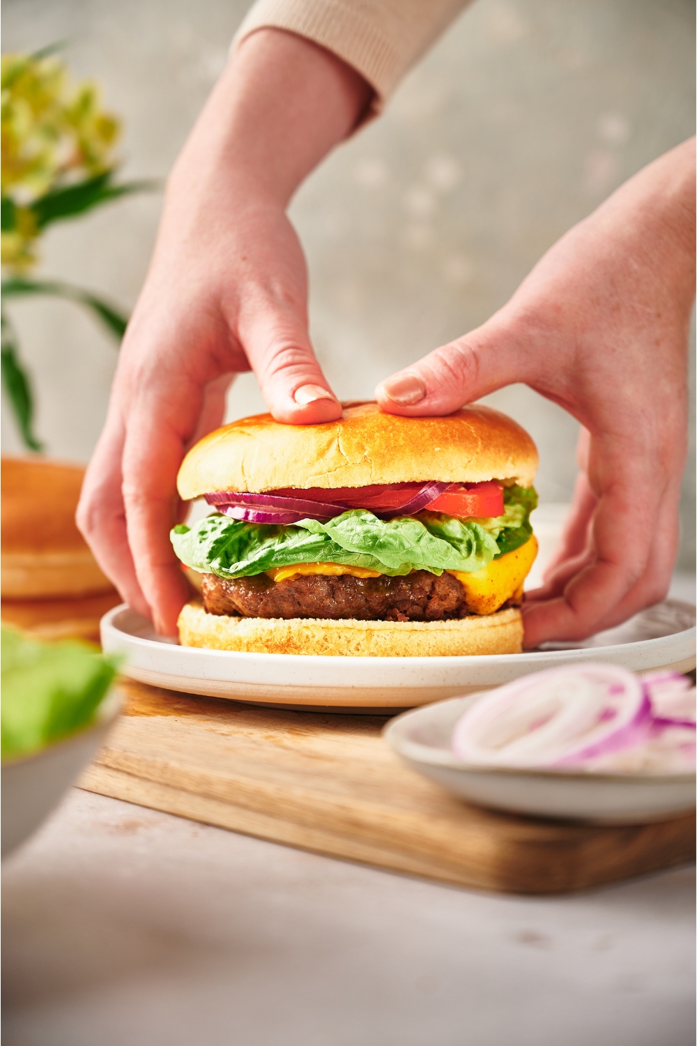 A hand grabbing a cheeseburger from a white plate. The burger has lettuce, red onion, sliced tomato, and cheese on it.